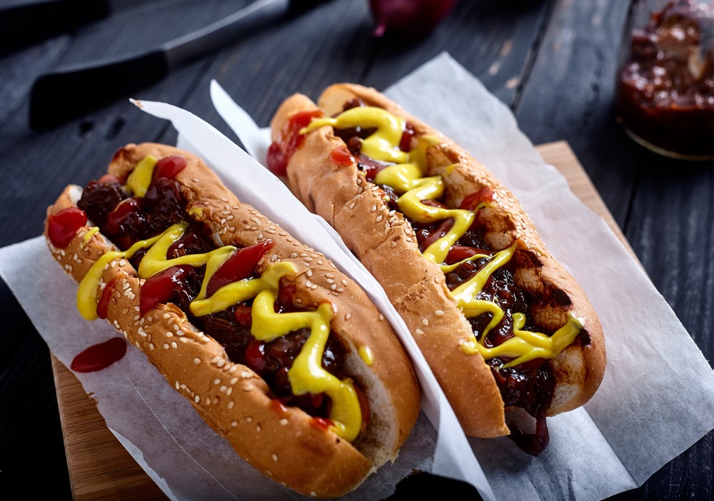 Homemade New York Style Hot dog. Kiełbasa z sosem cebulowym, ketchup i musztarda w białej bułce. Drewniane tło z cebulą, sosem i szczypcami na plecach