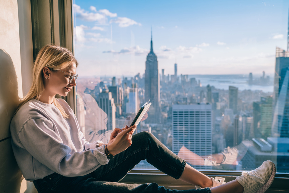 Attractive blonde hair female reading electronic book on digital tablet while sitting on hotel window sill with scenery view of Empire State building and New York downtown. Hipster girl travel blogger