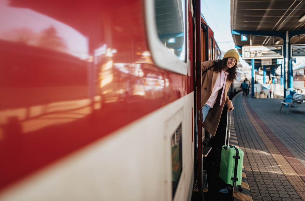 Szczęśliwa młoda podróżniczka z bagażem wysiadająca z pociągu na peronie stacji kolejowej, licencja: shutterstock/By Halfpoint